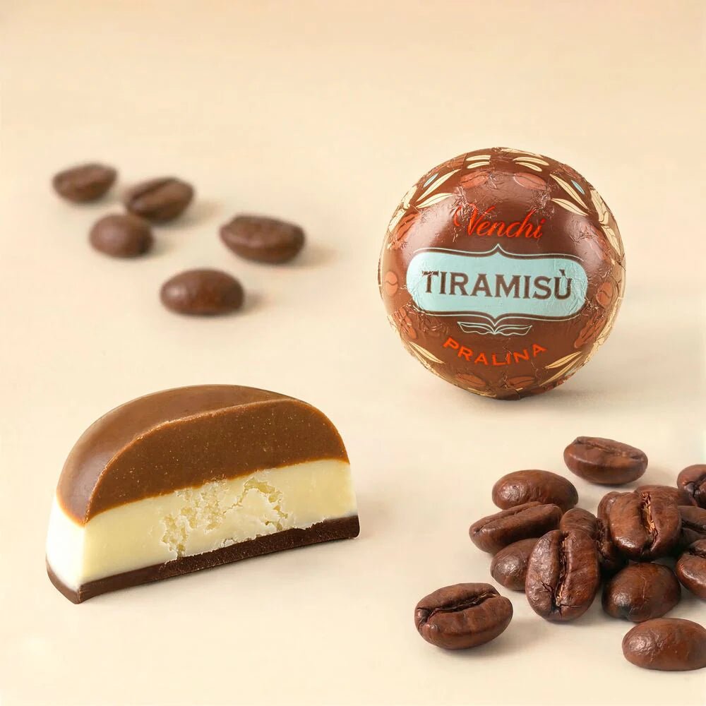 A round chocolate pralina wrapped in branded foil next to a cut half-chocolate showing a creamy off-white center, with scattered coffee beans on a warm beige background. The brand name 'Venchi TIRAMISU' is visible on the wrapper.
