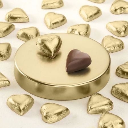 A round gold-toned chocolate tin or display stand on a white background, with two heart-shaped chocolates placed on top: one wrapped in shiny gold foil and one unwrapped in dark brown chocolate. More heart-shaped chocolates are scattered around the stand.
