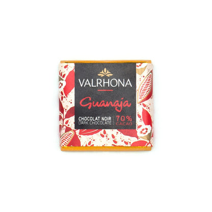 A single Valrhona Guanaja chocolate napolitain wrapped in a cream-colored paper sleeve with red and gold cacao pod illustrations and a black center label, photographed on a plain white background.