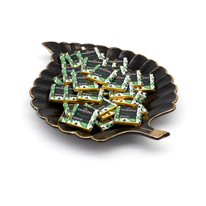 A pile of individually wrapped Valrhona Manjari napolitains on a decorative black leaf-shaped plate with gold trim, set against a clean white background.