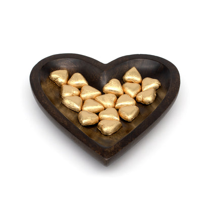 A heart-shaped dark wooden bowl filled with small gold foil-wrapped chocolates, photographed on a clean white background.